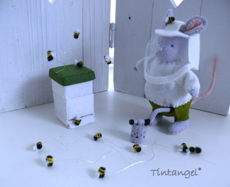 In this picture: a handmade felt mouse dressed as a beekeeper, with white jacket, bee hood, beehive, smoke pipe and many bees. All this is is made with PDF hand sewing pattern from WoolFeltWonders by Tintangel and is part of a series with felt toy animals and all kinds of clothing sets to mix and match to portray hobby or profession. Easy to make animals and clothing sets for decoration and play.