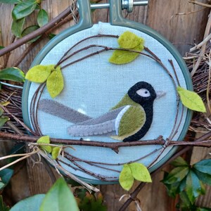 In this picture:  hand made wall decoration with a felt birds on linen in an embroidery hoop, the bird is a Great tit. The hoop is lying between  leaves in front of a wooden wall. This easy to make wall decoration is made with a PDF hand sewing pattern from WoolFeltWonders by Tintangel.