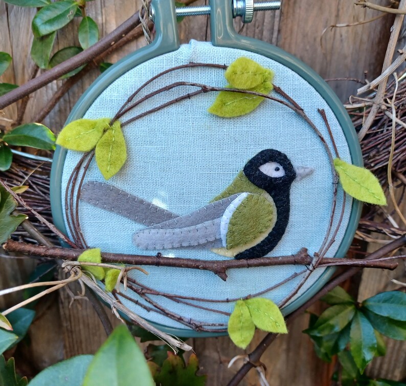 In this picture:  hand made wall decoration with a felt birds on linen in an embroidery hoop, the bird is a Great tit. The hoop is lying between  leaves in front of a wooden wall. This easy to make wall decoration is made with a PDF hand sewing pattern from WoolFeltWonders by Tintangel.