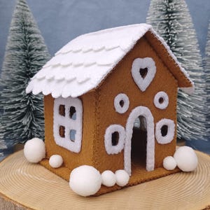 May include: A felt gingerbread house with a white scalloped roof and white accents. The house has a heart-shaped window and round and rectangular windows. White snowballs are at the base of the house. The house is on a wooden surface with miniature Christmas trees in the background.