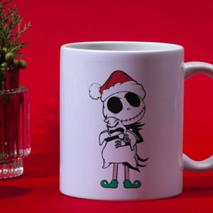 Puede incluir: Taza de cer&aacute;mica blanca con un dise&ntilde;o navide&ntilde;o con Jack Skellington de Pesadilla antes de Navidad con un gorro de Pap&aacute; Noel y sosteniendo un fantasma blanco. La taza tiene un asa negra.