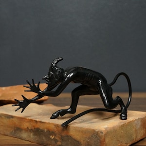 May include: A black metal figurine of a crouching creature with a pointed head, long limbs, and a thin tail. The figure has sharp facial features, extended fingers, and hooved feet, captured in a dynamic, low-to-the-ground pose on a flat surface.