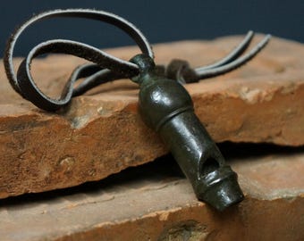 May include: A dark green metal whistle with a leather cord, resting on two weathered, reddish-brown bricks. The whistle has a cylindrical body with a small opening and a loop for the cord. The leather cord is dark brown.