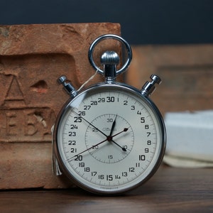 May include: A silver stopwatch with a white face and black hands. The stopwatch has a large outer ring with numbers 1-30 and a smaller inner ring with numbers 5-30. The stopwatch is sitting on a wooden surface.