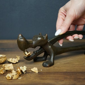 Può includere: Un schiaccianoci di bronzo a forma di lupo con una noce rotta nella sua bocca. Lo schiaccianoci è su una superficie di legno con pezzi di guscio di noce sparsi intorno.