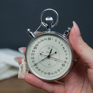 May include: A silver stopwatch with a white face and black hands. The stopwatch has a large outer ring with numbers 1-30 and a smaller inner ring with numbers 5-30. The stopwatch is being held in a hand.