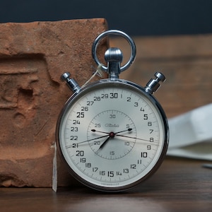 May include: A silver stopwatch with a white face and black hands. The stopwatch has a large dial with numbers from 1 to 30 and a smaller dial with numbers from 5 to 60. The stopwatch is set to 25 seconds.