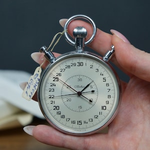 Könnte beinhalten: Eine silberne Stoppuhr mit weißem Zifferblatt und schwarzen Markierungen. Die Stoppuhr hat ein großes Zifferblatt mit den Zahlen von 1 bis 30 und ein kleineres Zifferblatt mit den Zahlen von 5 bis 30. Die Stoppuhr ist auf 25 Sekunden eingestellt.