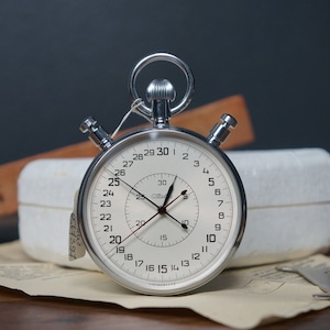 Könnte beinhalten: Eine silberne Stoppuhr mit weißem Zifferblatt und schwarzen Zeigern. Die Stoppuhr hat einen großen Ring um das Zifferblatt mit den Zahlen 1-30. Die Stoppuhr liegt auf einer weißen Box und einem Stück Papier.