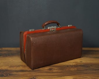 Op de afbeelding: Een vintage dokters tas van bruin leer met rode bies en een metalen sluiting.