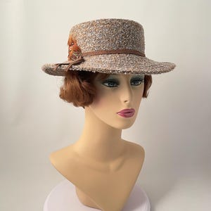 May include: A tweed-textured hat with a brown leather band and feather accents. The hat has a flat brim and a cylindrical crown. The hat is displayed on a mannequin head with a neutral skin tone and red hair.