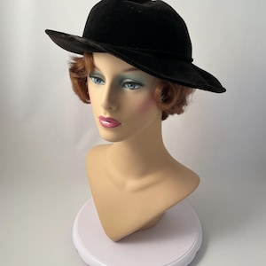 May include: A black fedora hat with a wide brim, displayed on a mannequin head. The hat is made of a soft, velvety material. The mannequin has fair skin, blue eyes, and red hair.
