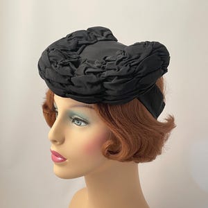 May include: A black vintage hat with a unique, ruffled design. The hat is made of a dark fabric and features a rounded crown with a decorative, textured trim. The hat is displayed on a mannequin head with red hair and a fair complexion.