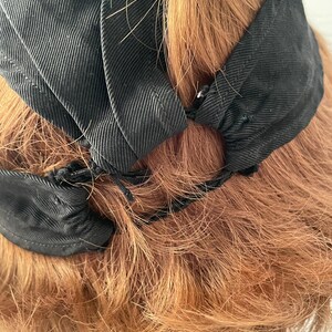 May include: Close-up of a black fabric head covering with a pleated design. The head covering is set against a backdrop of reddish-brown hair. The fabric appears to be a matte material, and the design includes small, gathered accents.