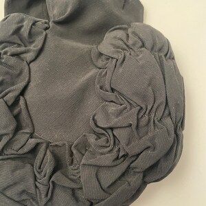 May include: A dark gray, textured fabric hat with a rounded crown and ruffled, gathered fabric details. The hat's material has a subtle striped pattern, and the overall design is soft and sculptural.