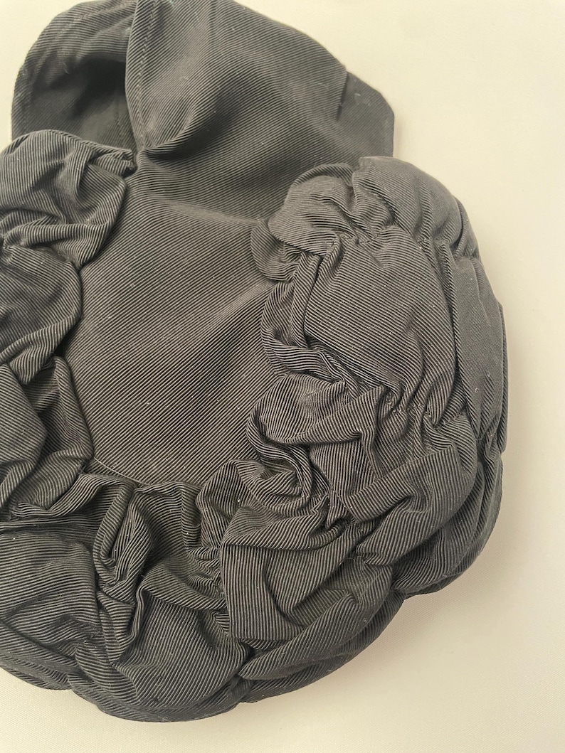 May include: A dark gray, textured fabric hat with a rounded crown and ruffled, gathered fabric details. The hat's material has a subtle striped pattern, and the overall design is soft and sculptural.