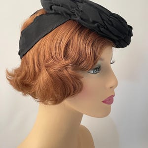 May include: A black vintage hat with a textured, pleated crown and a wide, smooth band. The hat is set against a neutral background, showcasing its classic design and elegant silhouette. This accessory is a timeless piece.