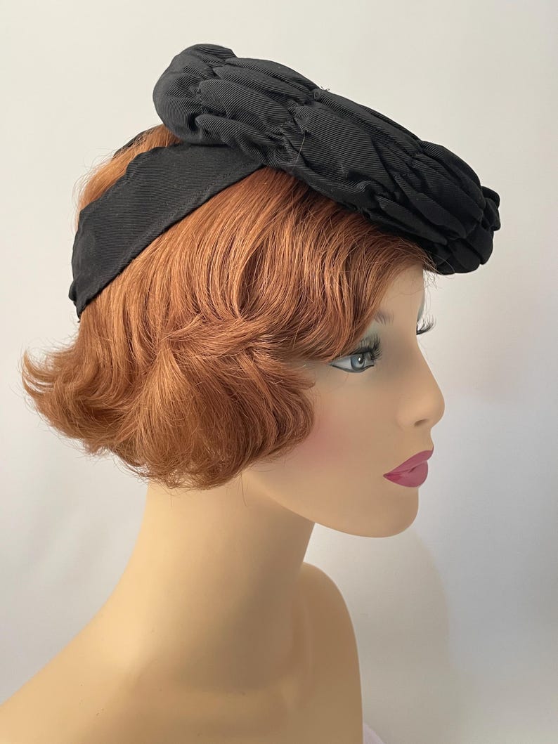 May include: A black vintage hat with a textured, pleated crown and a wide, smooth band. The hat is set against a neutral background, showcasing its classic design and elegant silhouette. This accessory is a timeless piece.