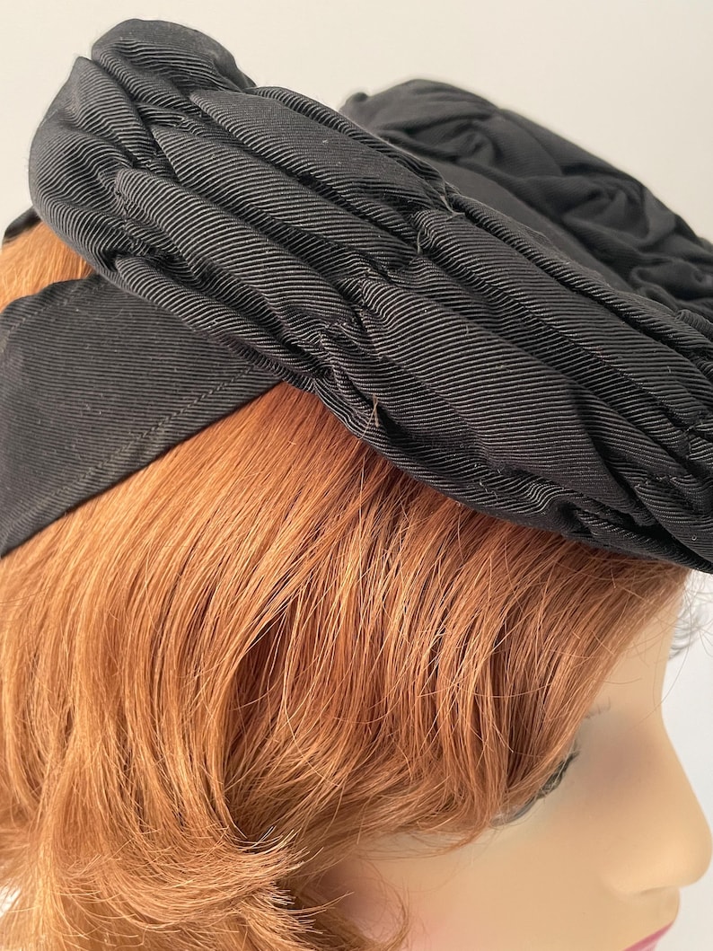 May include: A black hat with a pleated design. The hat has a wide band and a textured, ribbed fabric. The hat is worn by a person with red hair and fair skin. The hat appears to be vintage or retro.