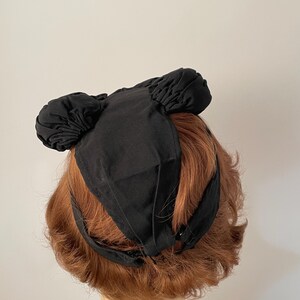 May include: A black fabric head covering with two rounded, gathered sections on top. The head covering has a band that wraps around the head and is secured with a button. The head covering is shown on a mannequin head with auburn hair.