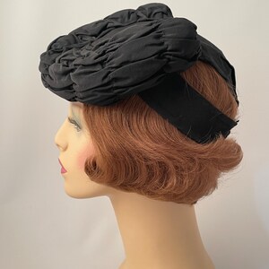 May include: A black hat with a gathered fabric design. The hat features a wide black band and is displayed on a mannequin head with auburn hair and a fair complexion. The hat's texture and shape are prominent.