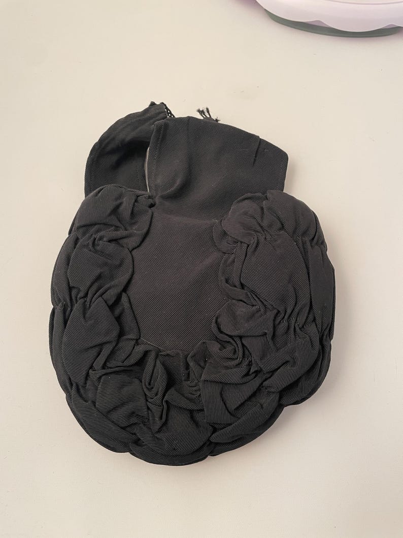 May include: Black, gathered fabric clutch with a rounded shape. The clutch features a ruched design around the edges and a structured top with a small handle. The fabric appears to be a ribbed material.