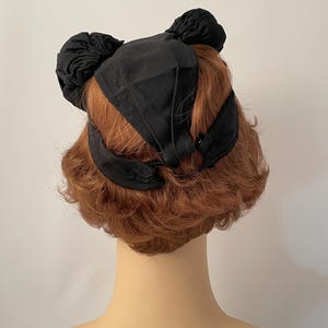 May include: A black hat with a unique design, featuring two bun-like accents on top and a ribbon detail. The hat is set against a neutral background, showcasing its shape and texture.