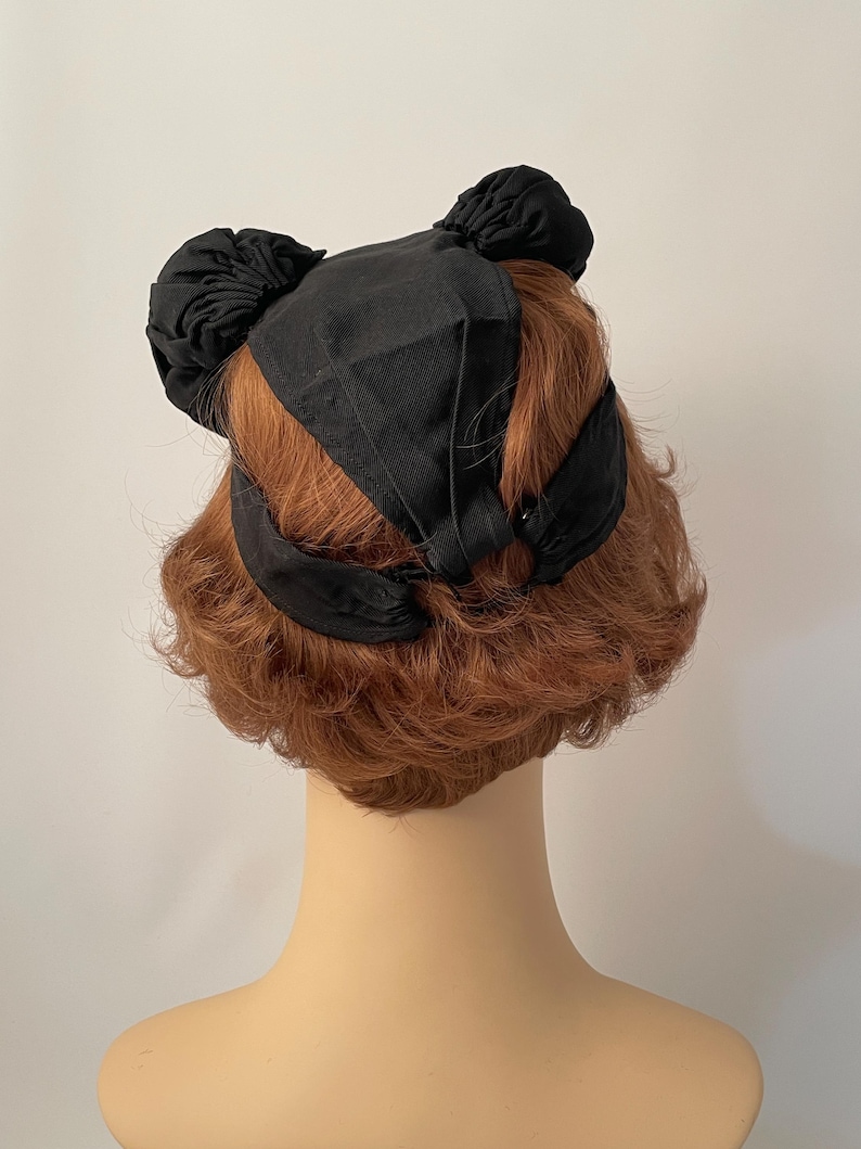 May include: A black hat with a unique design, featuring two bun-like accents on top and a ribbon detail. The hat is set against a neutral background, showcasing its shape and texture.