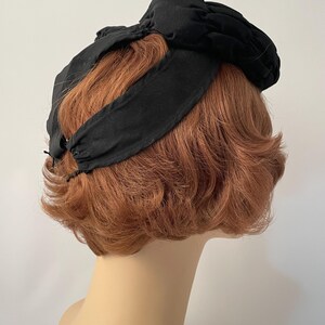 May include: A black vintage hat with a knotted bow detail. The hat features a wide headband and a gathered fabric crown. The hat is displayed on a mannequin head with short, auburn hair, against a plain white background.