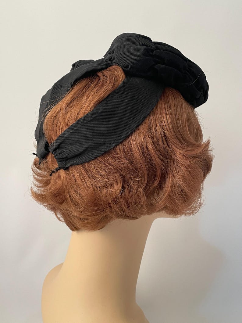 May include: A black vintage hat with a knotted bow detail. The hat features a wide headband and a gathered fabric crown. The hat is displayed on a mannequin head with short, auburn hair, against a plain white background.