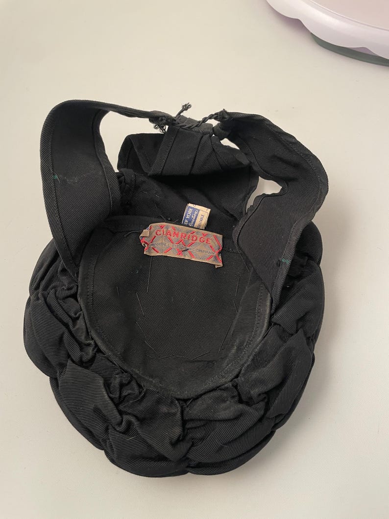 May include: A black hat with a unique design, featuring a rounded crown and a structured brim. The hat has a label that reads "CIANRIDGE". The hat's design includes a series of gathered fabric details, adding texture and visual interest. The hat is made of a dark fabric.