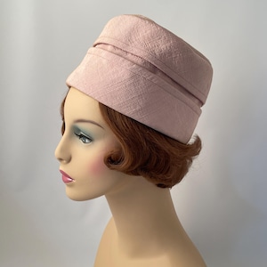 May include: A light pink, vintage-style hat with a textured weave. The hat has a structured, pillbox design with a slightly angled brim. The hat is displayed on a mannequin head with red hair and a fair complexion.