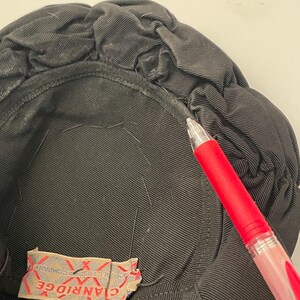 May include: Close-up of a black hat with a gathered, textured design. The interior is lined with black fabric. A red pen is in the frame, pointing towards the hat. A tag with the text "CLANRIDGE" is visible.