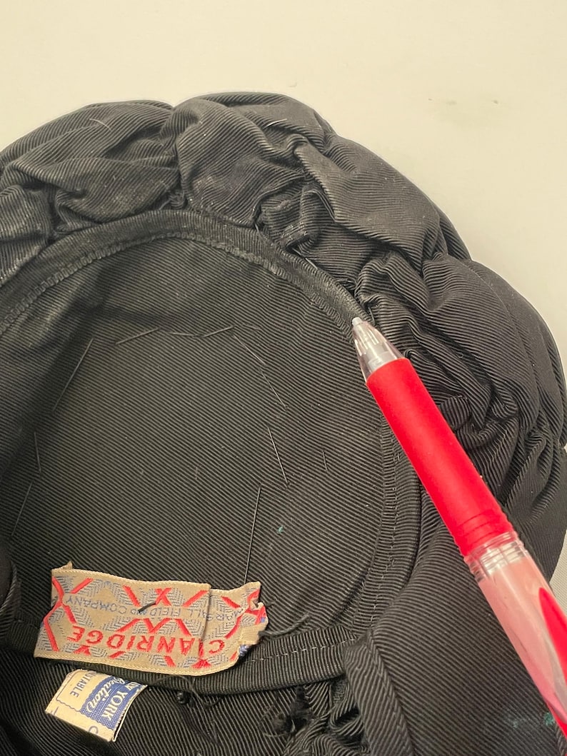 May include: Close-up of a black hat with a gathered, textured design. The interior is lined with black fabric. A red pen is in the frame, pointing towards the hat. A tag with the text "CLANRIDGE" is visible.