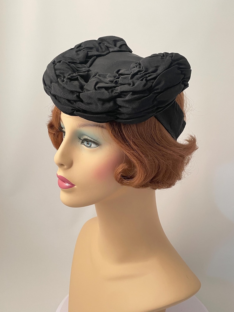 May include: A black vintage hat with a unique, ruffled design. The hat is made of a dark fabric and features a rounded crown with a decorative, textured trim. The hat is displayed on a mannequin head with red hair and a fair complexion.