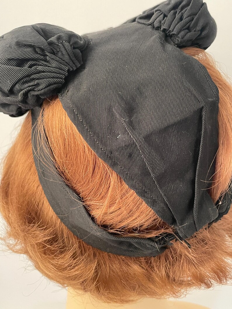 May include: A black headband with two decorative, gathered fabric accents. The headband is made of a ribbed fabric and is worn on a person's head with reddish-brown hair. The headband appears to be vintage or retro in style.