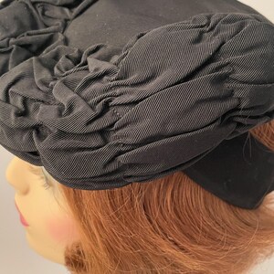 May include: A black hat with a rounded crown and a wide brim. The hat is made of a textured fabric, with gathered detailing around the crown and brim. The hat is a classic style, suitable for formal occasions.