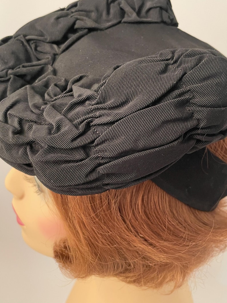 May include: A black hat with a rounded crown and a wide brim. The hat is made of a textured fabric, with gathered detailing around the crown and brim. The hat is a classic style, suitable for formal occasions.