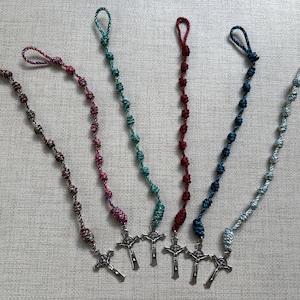 May include: Six colorful rosaries with knotted beads and silver crucifix pendants. The rosaries are in shades of brown, pink, teal, red, blue, and light blue. Each rosary has a loop at the top for hanging.