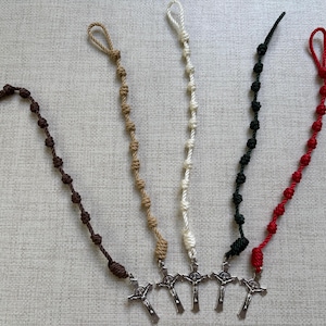 May include: Five rosaries in various colors: brown, tan, white, black, and red. Each rosary features knotted beads and a silver crucifix pendant. The rosaries are arranged on a textured, light-colored surface.