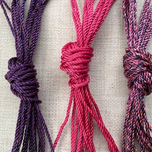 May include: Three spools of colourful rope, each tied in a knot. The rope on the left is purple with a dark blue stripe, the middle rope is pink, and the rope on the right is a mix of pink, purple, and blue.