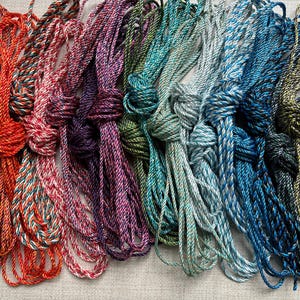 May include: A selection of multi-coloured twisted cords in shades of red, orange, pink, purple, green, blue, and yellow. Each cord is knotted at the top, with the strands spreading out below. These cords are likely for crafting or decorative use.