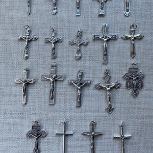 K&ouml;nnte beinhalten: Eine Sammlung von 19 silberfarbenen Kreuzanh&auml;ngern mit verschiedenen Designs, darunter einfache Kreuze, Kreuze mit Figuren und verzierte Kreuze.