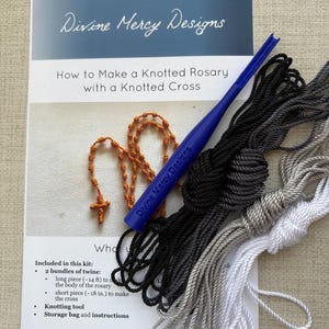 K&ouml;nnte beinhalten: Ein Kit zum Herstellen eines geknoteten Rosenkranzes mit einem geknoteten Kreuz. Das Kit enth&auml;lt zwei B&uuml;ndel Schnur, ein Knotenwerkzeug, eine Aufbewahrungstasche und Anweisungen. Die Schnur ist in Schwarz, Grau und Wei&szlig;. Die Anweisungen sind auf einer wei&szlig;en Karte mit dem Text "Divine Mercy Designs" und "Wie man einen geknoteten Rosenkranz mit einem geknoteten Kreuz herstellt" gedruckt.