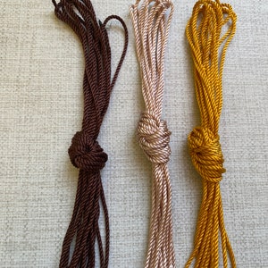 May include: Three spools of cord, one brown, one beige, and one gold, are tied in knots. The cords are arranged vertically against a white textured background.