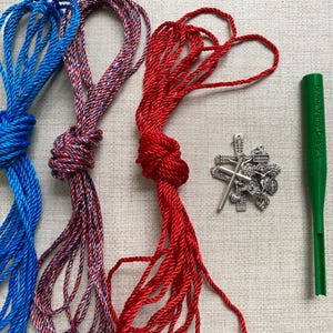 May include: Three knotted cords in blue, red-white-blue, and red. A silver crucifix pendant and a green tool are also present. The green tool has text printed on it. The items are arranged on a light beige surface.