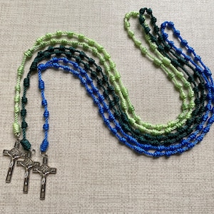 May include: Several rosaries in shades of green and blue are displayed. Each rosary features knotted cord and a silver crucifix pendant. The rosaries are arranged on a neutral-colored surface.