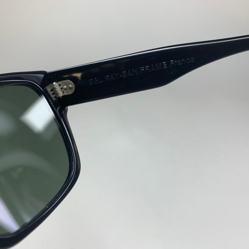 Peut inclure: Lunettes de soleil Ray-Ban &agrave; monture noire avec verres verts. Les lunettes ont une charni&egrave;re en m&eacute;tal et le texte "B&L RAY-BAN FRAME  F12108" est imprim&eacute; sur la monture.