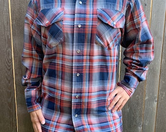 Può includere: Una camicia a quadri a maniche lunghe con un motivo blu, rosso e bianco. La camicia ha una chiusura con bottoni sul davanti e due tasche sul petto.