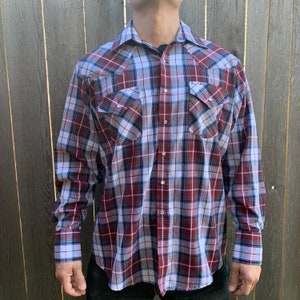 May include: A long-sleeved plaid shirt with a red, white, and blue pattern. The shirt has a button-down collar and two chest pockets.